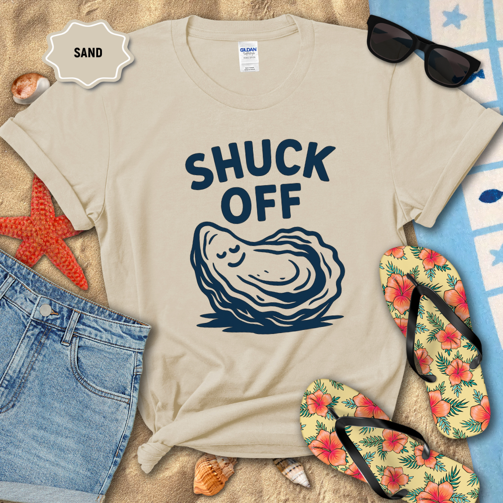 Beige t-shirt with oyster graphic and 'SHUCK OFF' text on a sandy background with flip-flops and sunglasses.