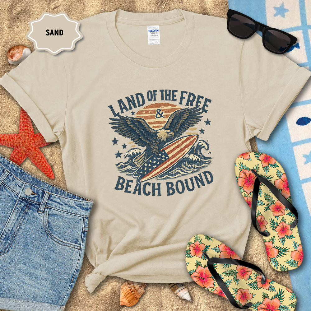 Beige t-shirt with 'Land of the Free & Beach Bound' design on sand with beach accessories.