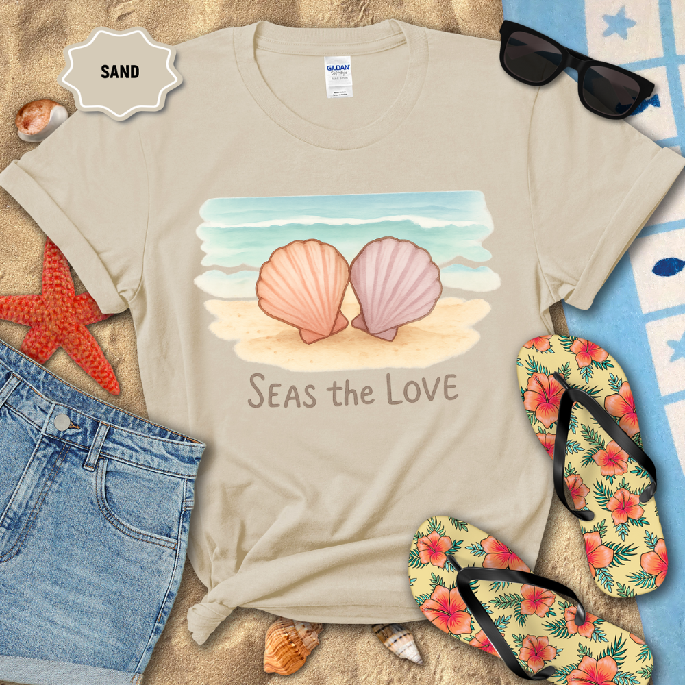 Beige t-shirt with shell design and 'Seas the Love' text on a sandy background with beach accessories.