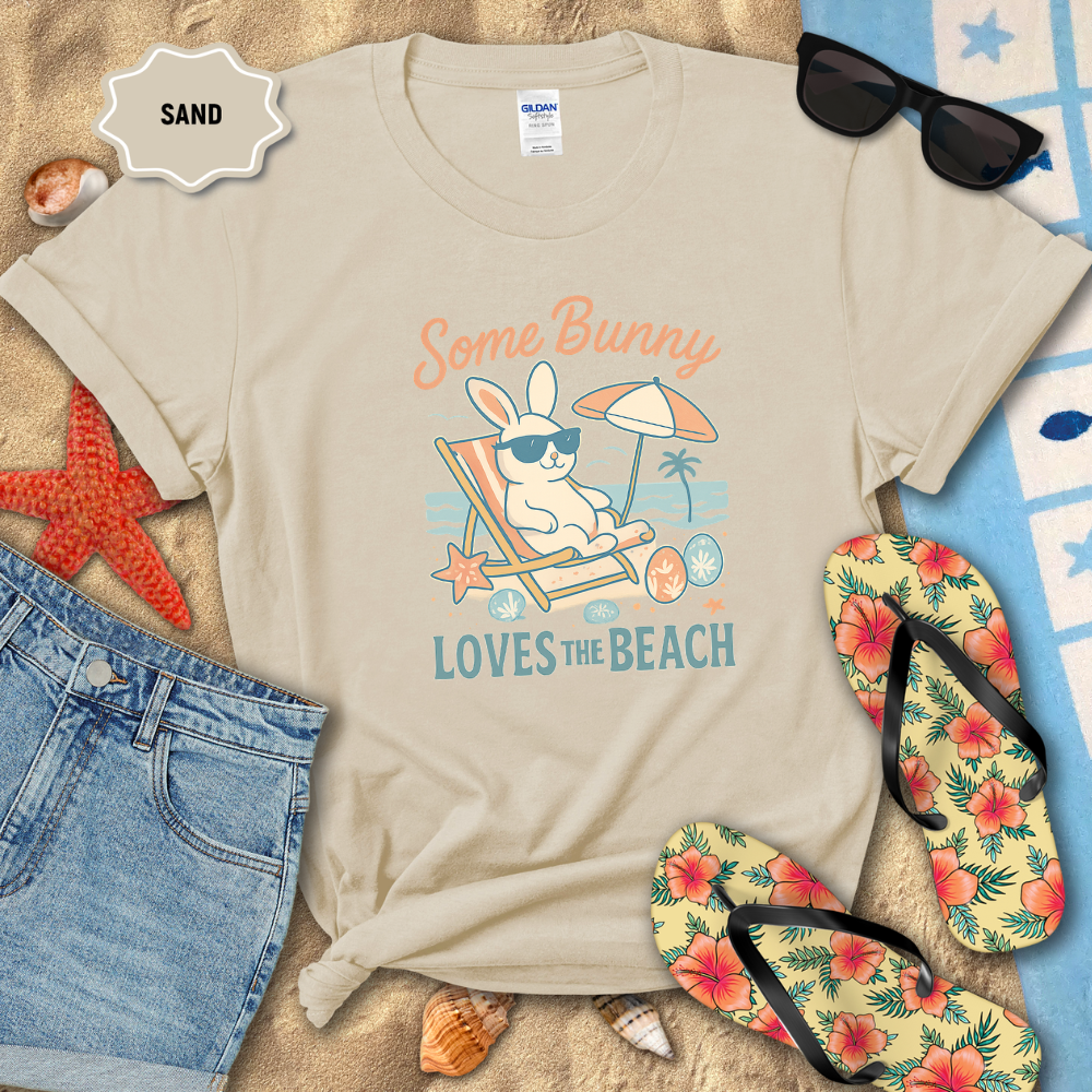 Beige t-shirt with bunny graphic and text 'Some Bunny Loves the Beach' on sand with beach accessories.