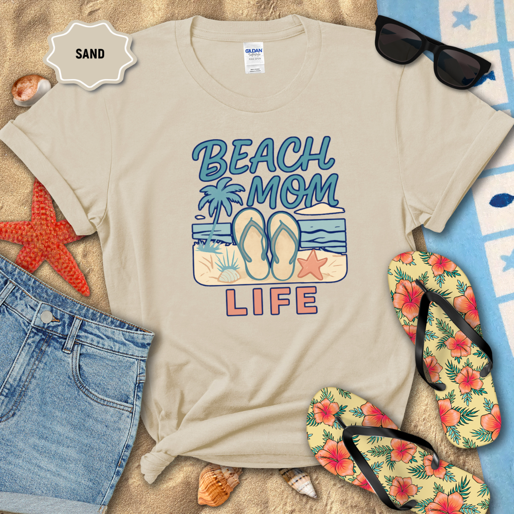 Beige t-shirt with 'Beach Mom Life' design on sand with flip-flops and sunglasses.