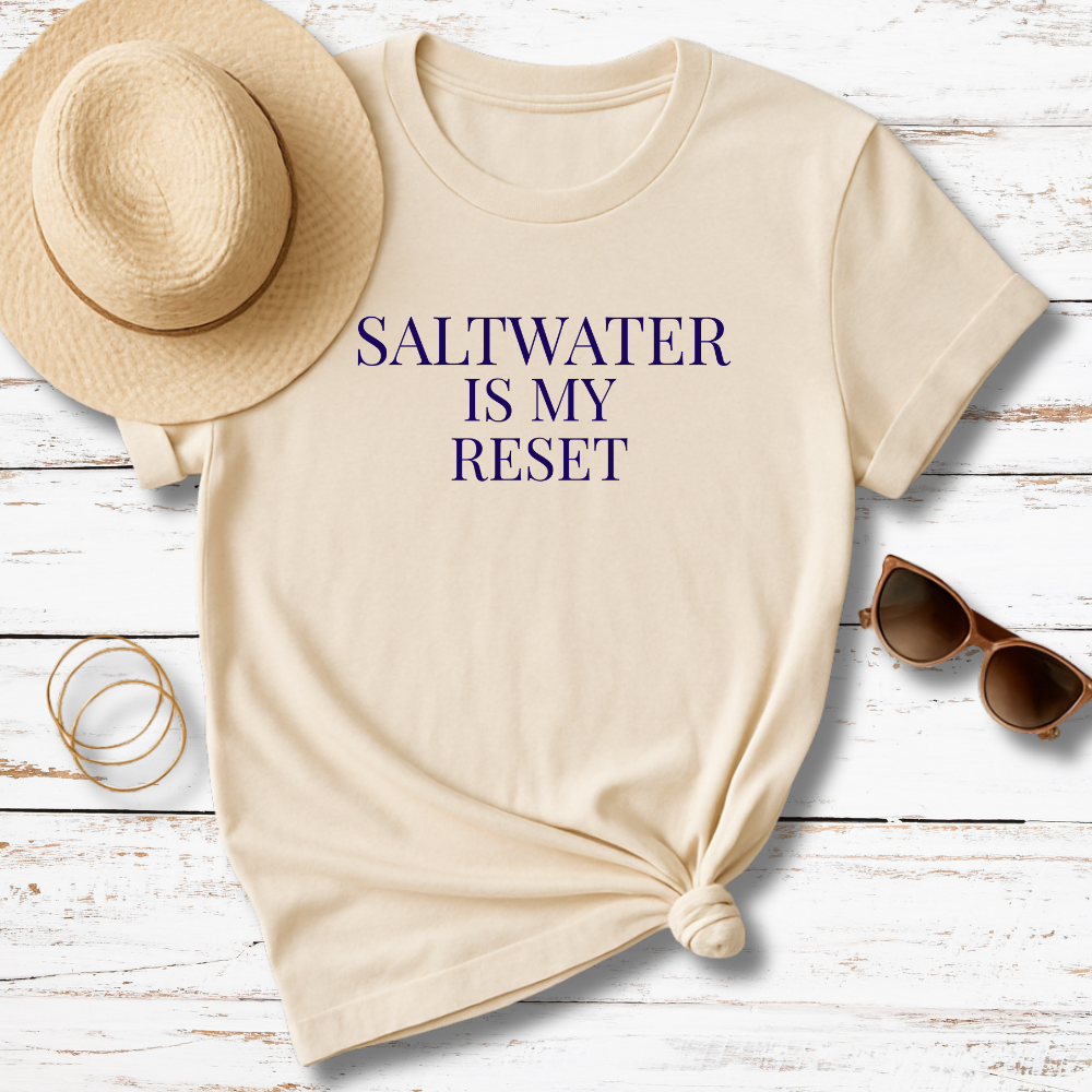 Beige t-shirt with 'SALTWATER IS MY RESET' text, sun hat, sunglasses, and bracelet on a wooden surface.