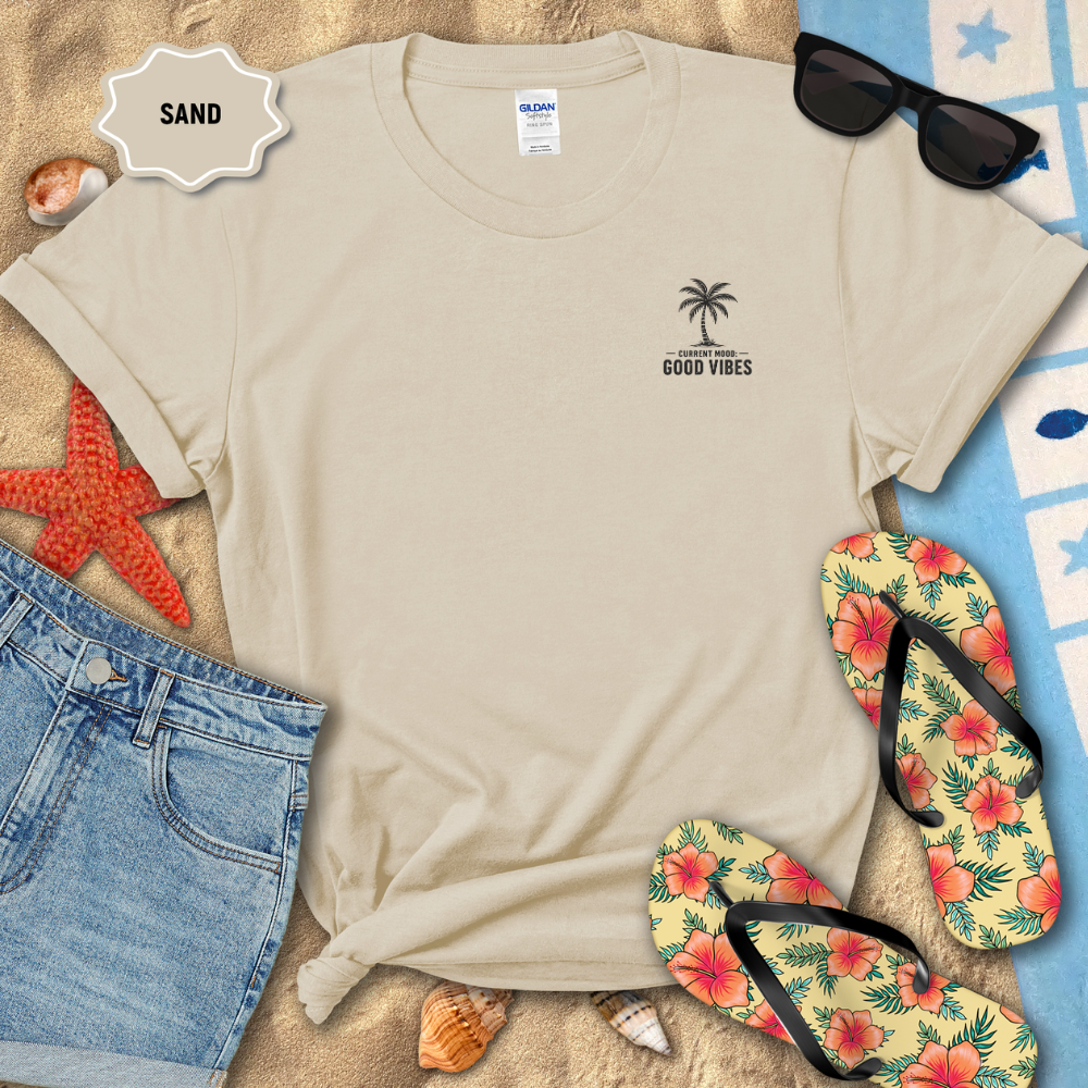 Beige t-shirt with palm tree design on sand with flip-flops and sunglasses.