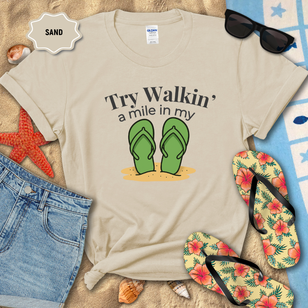Beige t-shirt with flip-flop graphic and text on a sandy background with sunglasses and flip-flops.