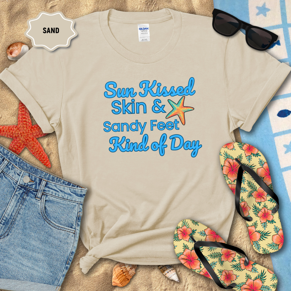 Beige t-shirt with 'Sun Kissed Skin & Sandy Feet Kind of Day' text, sunglasses, flip-flops, and beach items on sand.