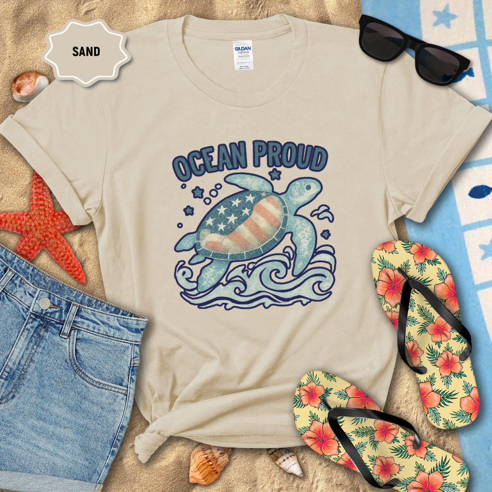 Beige t-shirt with turtle graphic and 'Ocean Proud' text on sand with beach accessories.