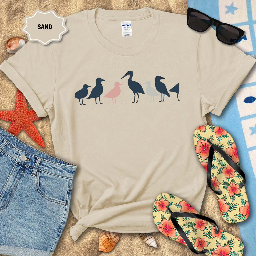 Beige t-shirt with seagull design on sand with beach accessories.
