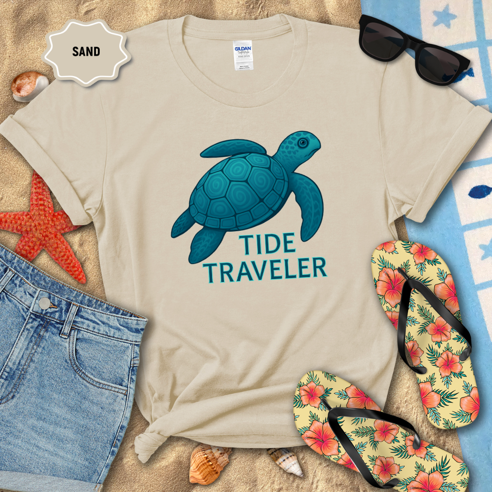 Beige t-shirt with blue turtle graphic and 'Tide Traveler' text, surrounded by beach accessories on sand.