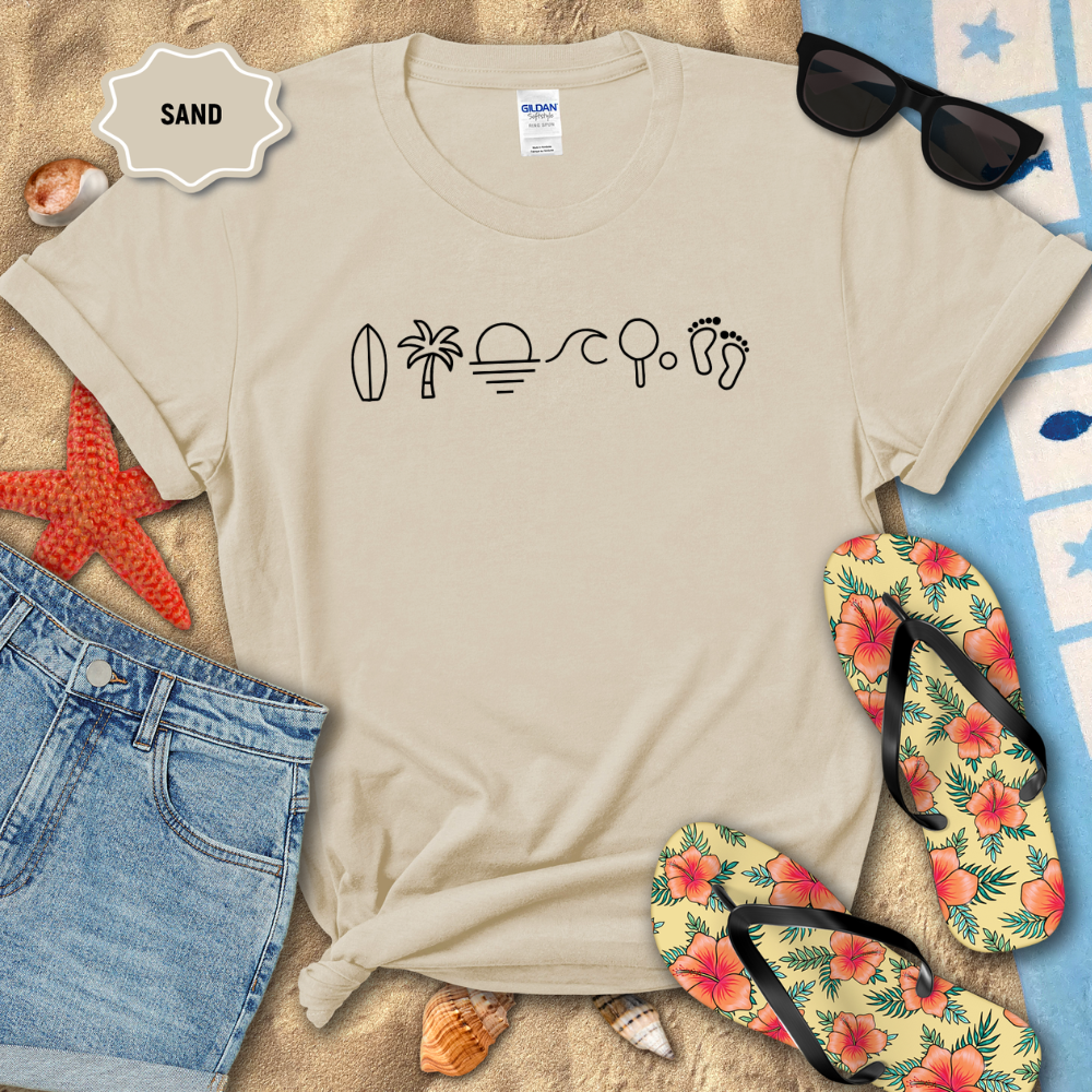 Beige t-shirt with beach-themed graphics and text, surrounded by beach accessories on sand.