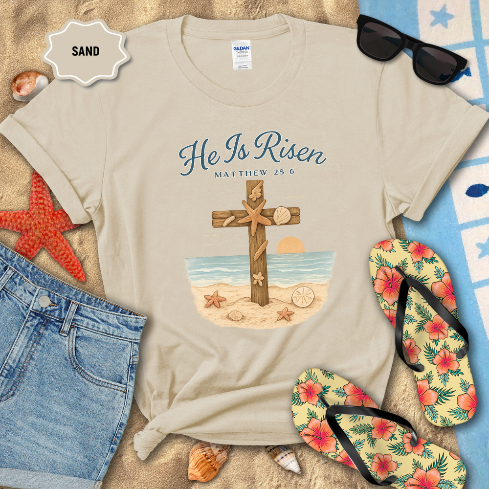 Beige t-shirt with 'He Is Risen' design on a sandy background with beach accessories.