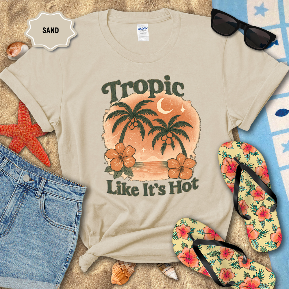 Beige t-shirt with tropical design and text 'Tropic Like It's Hot' on a sandy background with beach accessories.