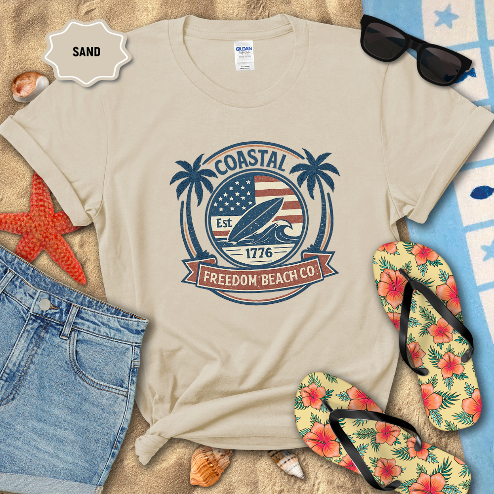 Beige t-shirt with 'Coastal Freedom Beach Co' design on sand with beach accessories.
