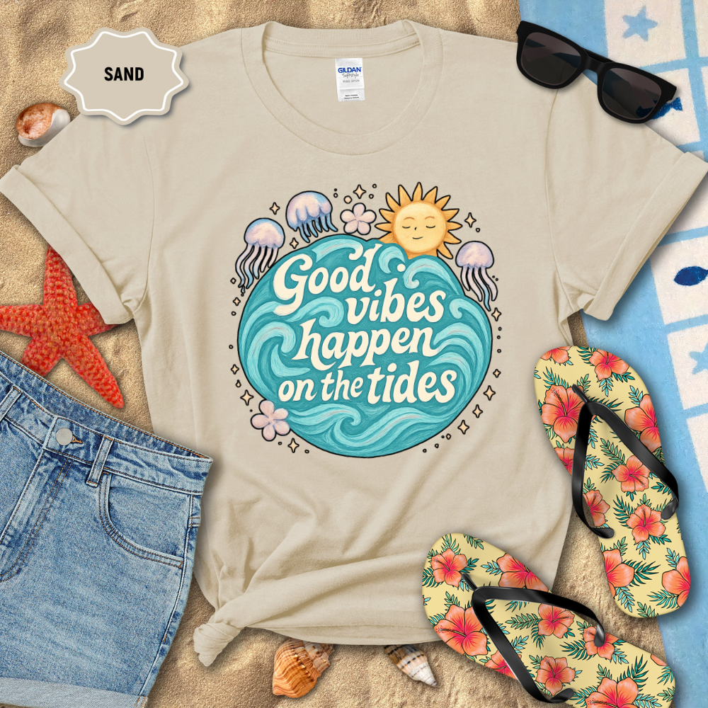 Beige t-shirt with 'Good vibes happen on the tides' design on sand with beach accessories.
