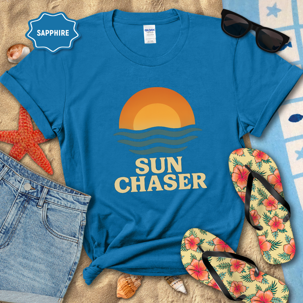 Blue t-shirt with 'Sun Chaser' design on a beach-themed background with sunglasses, flip-flops, and denim shorts.