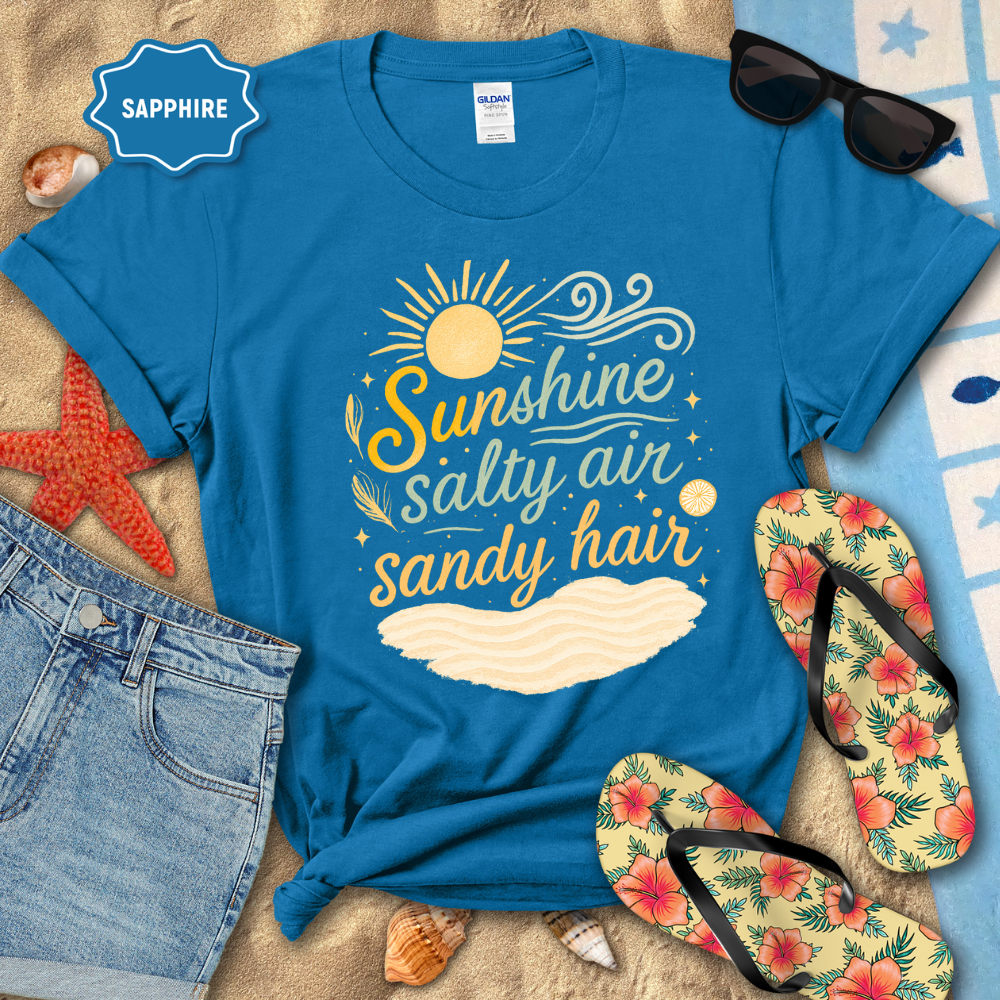 Blue t-shirt with 'Sunshine, Salty Air, Sandy Hair' design on a beach-themed background with sunglasses, flip-flops, and denim shorts.