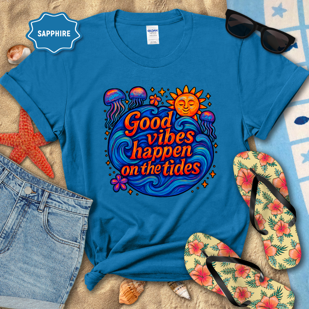 Blue t-shirt with 'Good vibes happen on the tides' design, sunglasses, flip-flops, and beach items on sand.