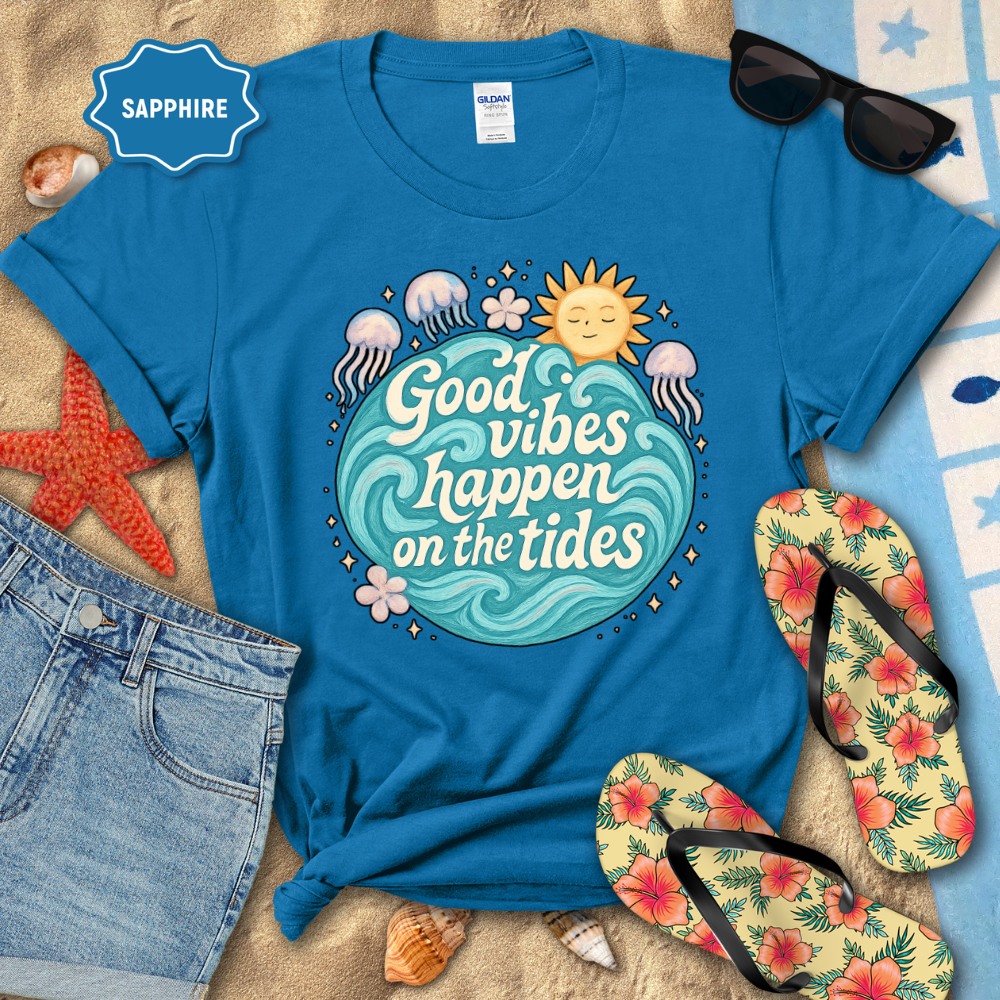 Blue t-shirt with 'Good vibes happen on the tides' design, sunglasses, flip-flops, and beach items on sand.