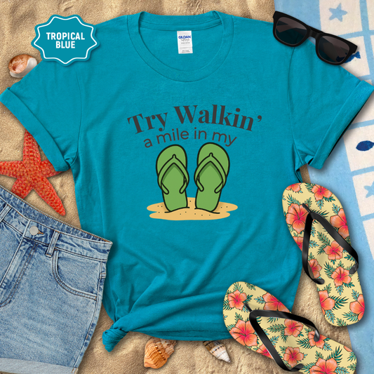 Blue t-shirt with flip-flop graphic and text, surrounded by beach accessories on a sandy background.