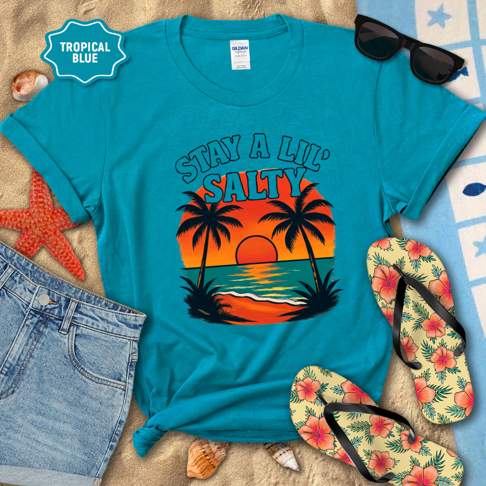 Blue t-shirt with 'Stay a Lil' Salty' design on a sandy background with flip-flops and sunglasses.
