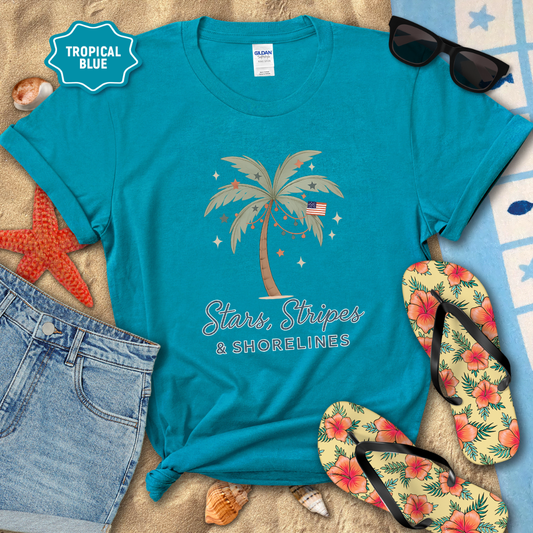 Blue t-shirt with palm tree design and text, surrounded by beach accessories on sand.