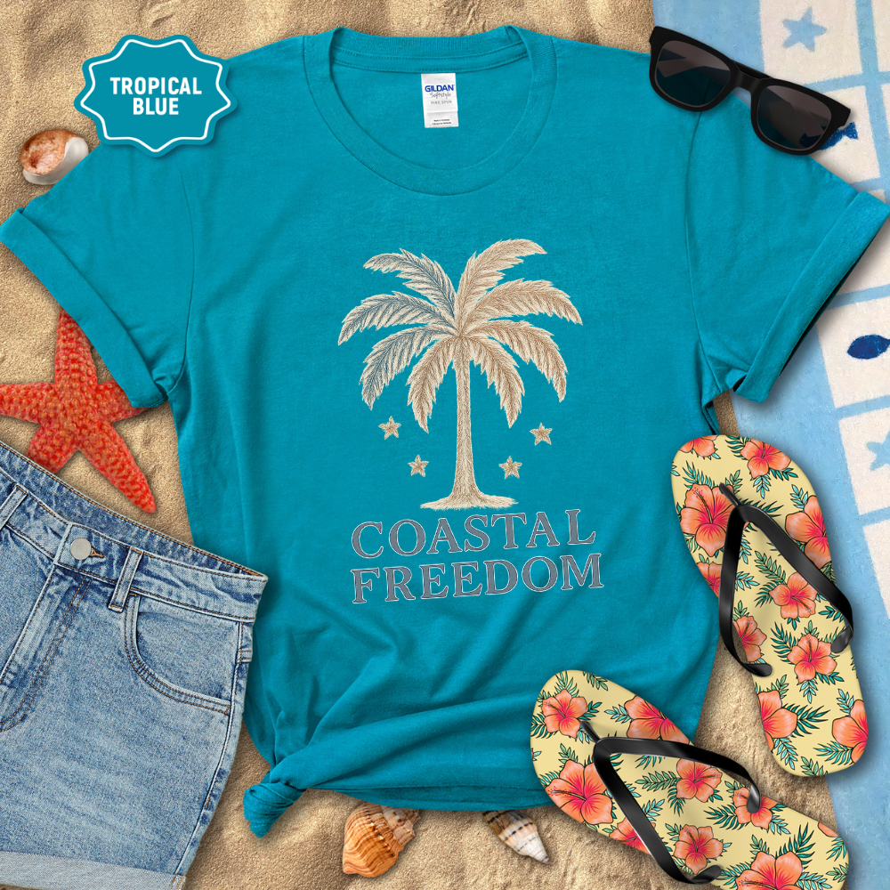 Blue t-shirt with palm tree design and 'Coastal Freedom' text, surrounded by beach accessories on sand.