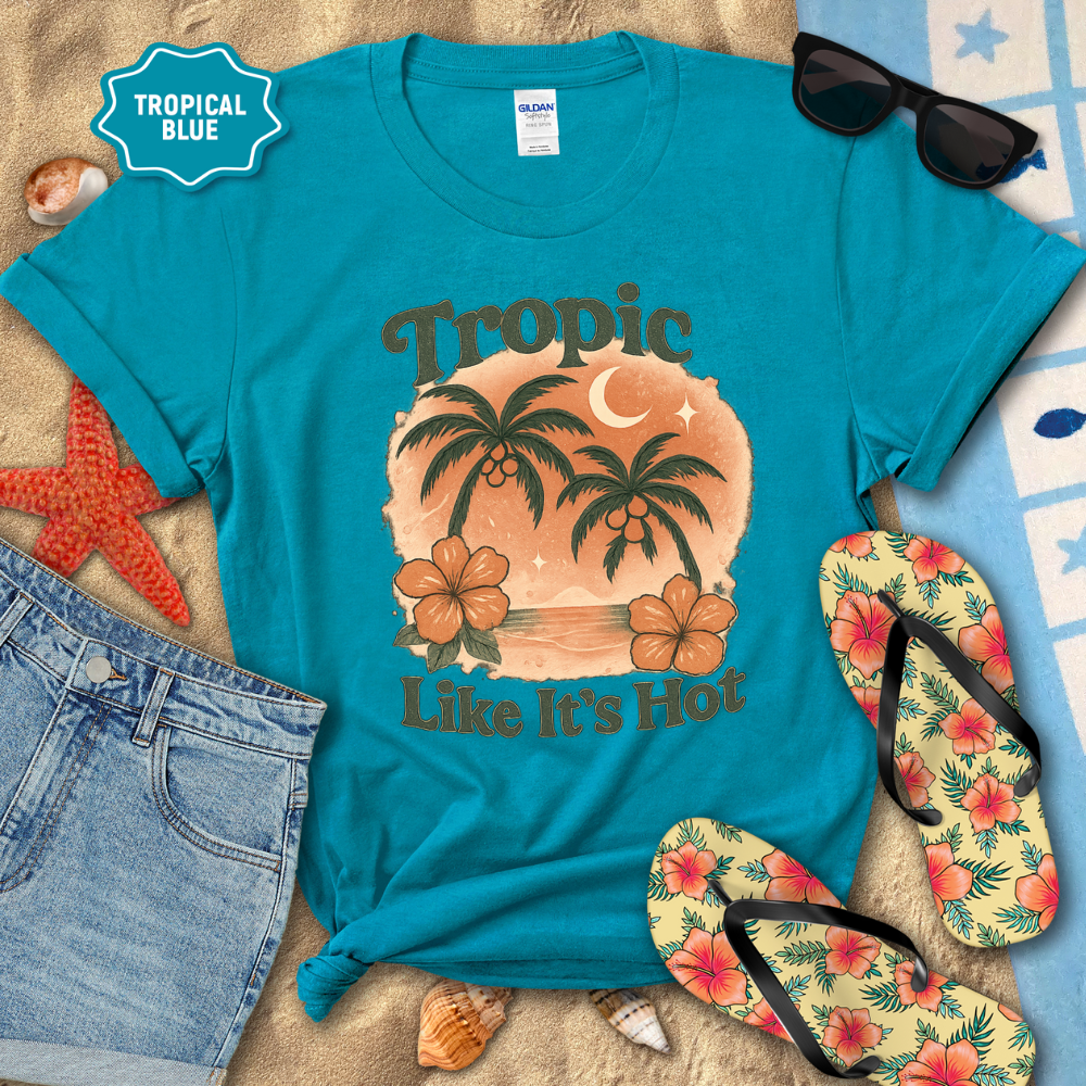Turquoise t-shirt with tropical design and text, surrounded by beach accessories on a sandy background.