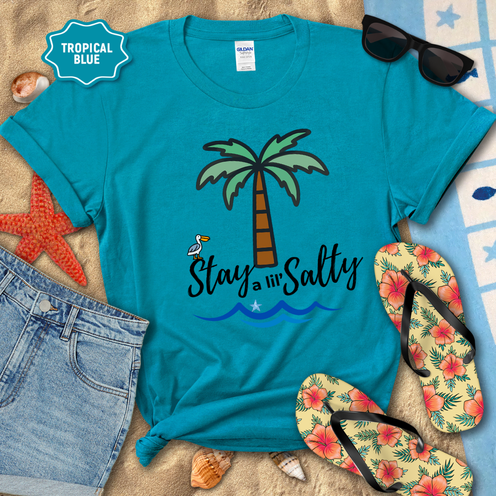 Blue t-shirt with palm tree and 'Stay a lil' Salty' text, surrounded by beach accessories on sand.