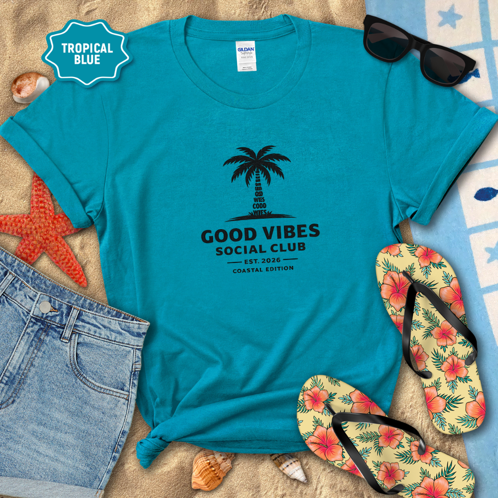 Blue t-shirt with palm tree design and 'Good Vibes Social Club' text, surrounded by beach accessories on sand.