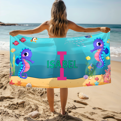 A person standing on a beach holding a personalized beach towel with an under the sea design, including seahorses and the name 'ISABEL' in large letters.