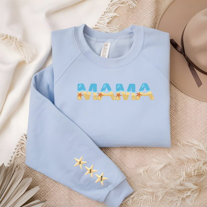Soft light blue crewneck featuring MAMA in sand and ocean tones with matching starfish down the sleeve, displayed as a flat ay on soft pink and white blankets with a hat in the background.