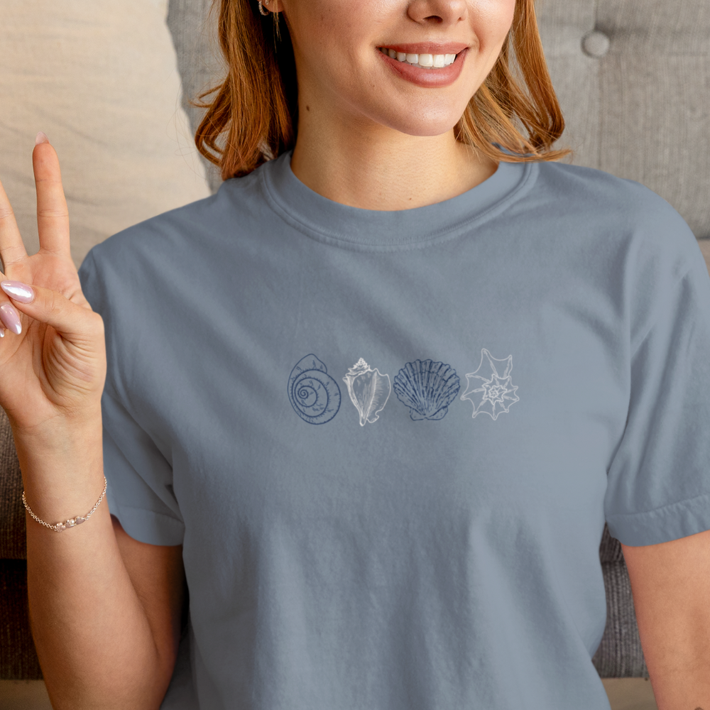 Smiling woman wearing a blue Comfort Colors t-shirt with minimalist coastal shell design, making a peace sign.