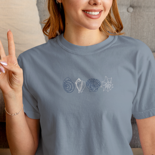 Smiling woman wearing a blue Comfort Colors t-shirt with minimalist coastal shell design, making a peace sign.