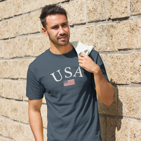 Man wearing a gray t-shirt with 'USA' and an American flag design, standing against a stone wall.