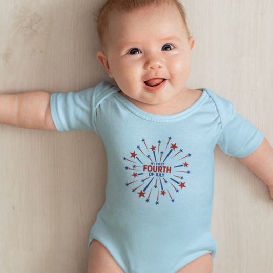 Smiling baby in a light blue “My First Fourth of July” bodysuit with red and blue firework design, lying on a light wood floor.