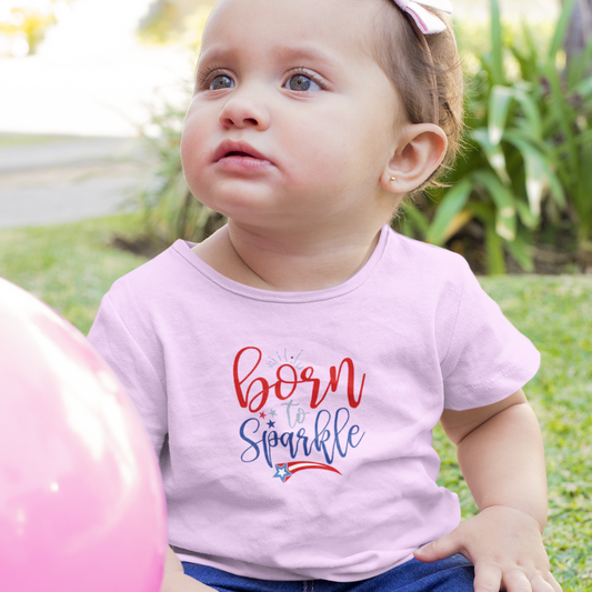Toddler girl sitting outside next to a pink balloon wearing a Pink kids' T-shirt with the phrase "Born to Sparkle" in red and blue lettering, accented with patriotic stars and a shooting star