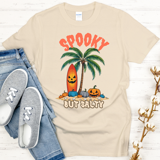 Beige t-shirt with Halloween-themed design featuring pumpkins, palm trees, and surfboard on a white wooden surface.