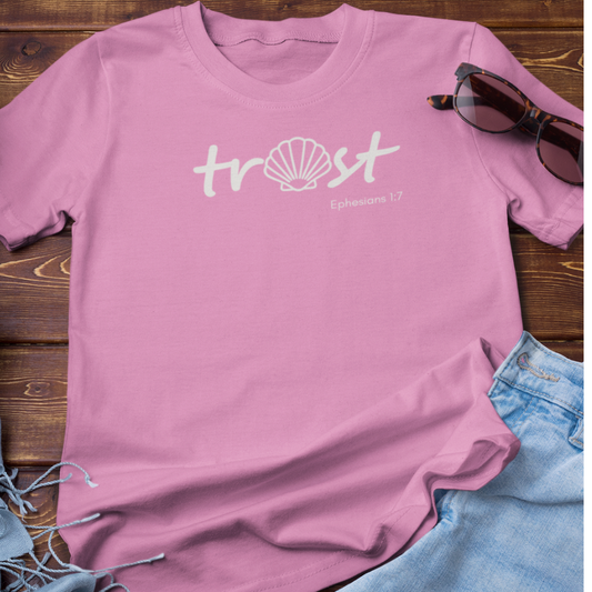 Pink t-shirt with 'trust' and shell design on a wooden surface with sunglasses and jeans.