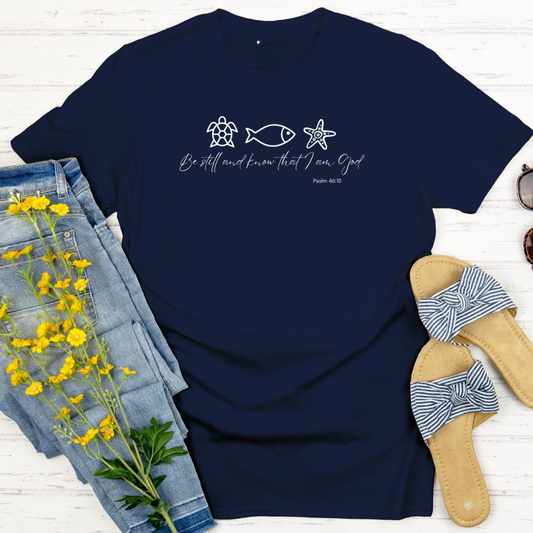 Navy blue t-shirt with a design and text, placed on a white wooden surface with denim shorts and sandals.