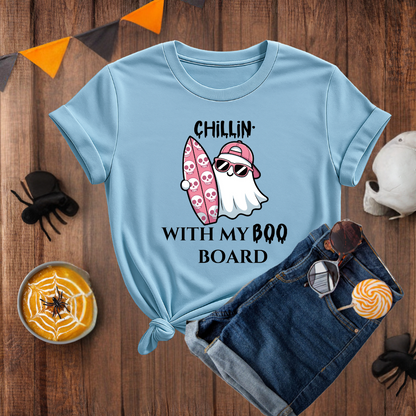 Light blue t-shirt with ghost and surfboard design on a wooden surface with Halloween decorations.