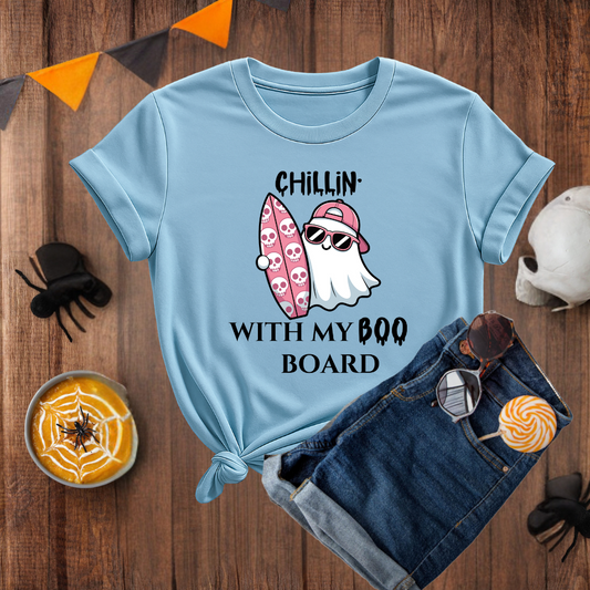 Light blue t-shirt with ghost and surfboard design on a wooden surface with Halloween decorations.