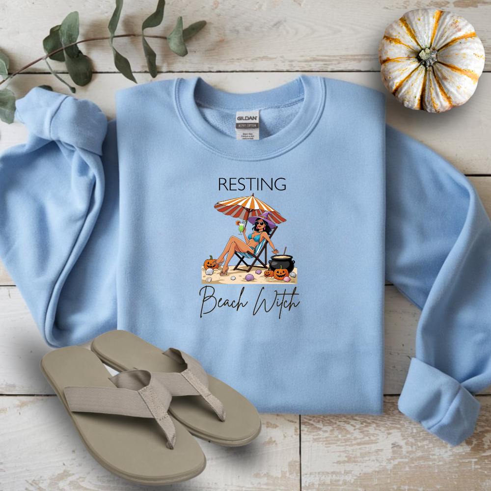 Light blue sweatshirt with 'Resting Beach Witch' design on a wooden surface with flip-flops and a small pumpkin.
