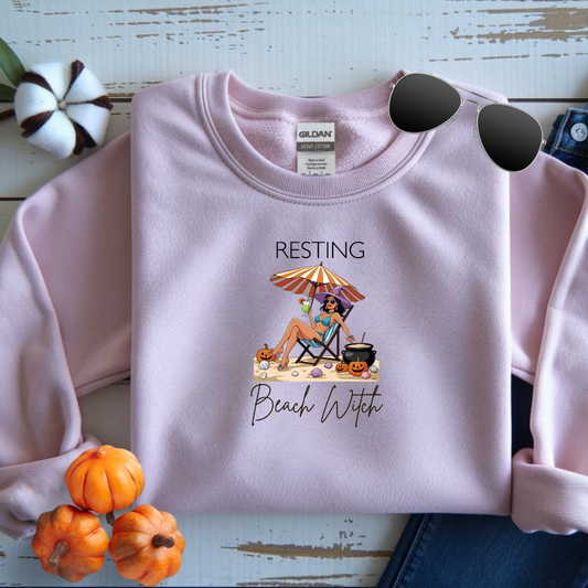 Pink sweatshirt with 'Resting Beach Witch' graphic on a wooden surface with pumpkins.