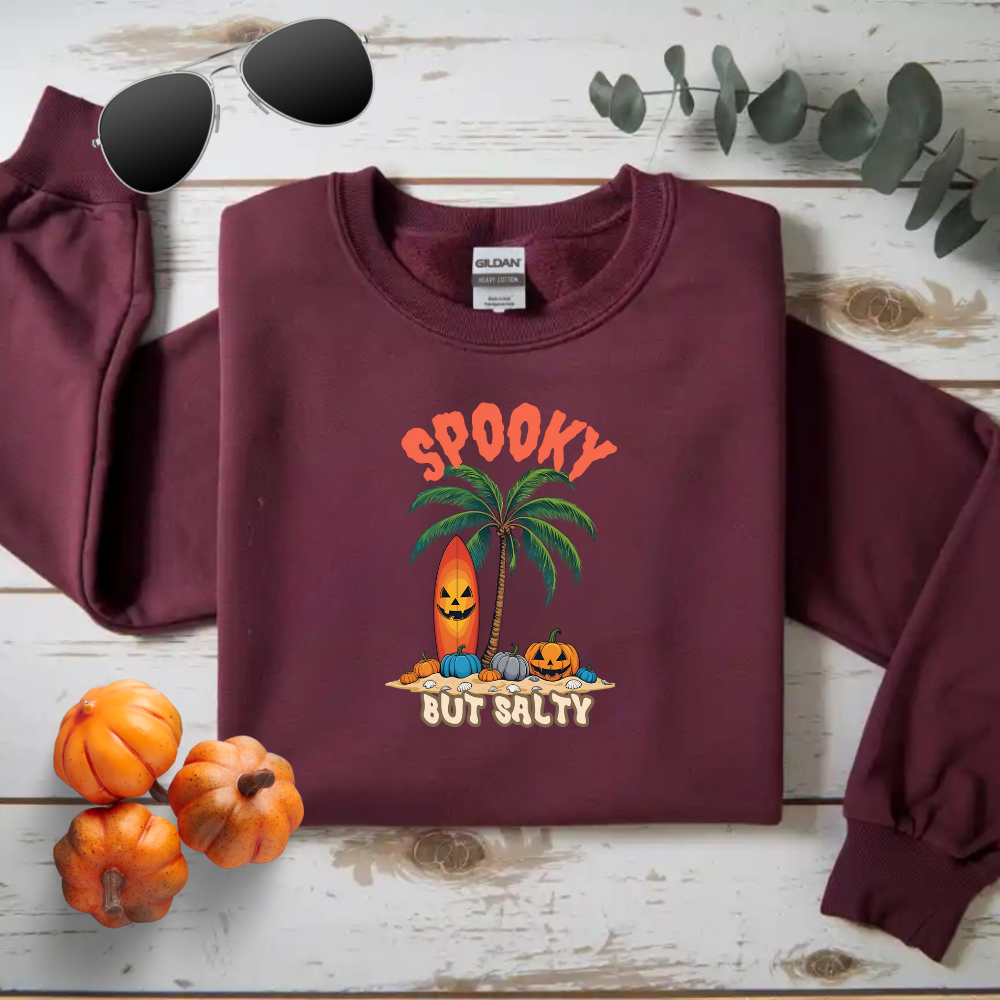 Maroon sweatshirt with Halloween-themed design on a wooden surface with pumpkins and sunglasses.