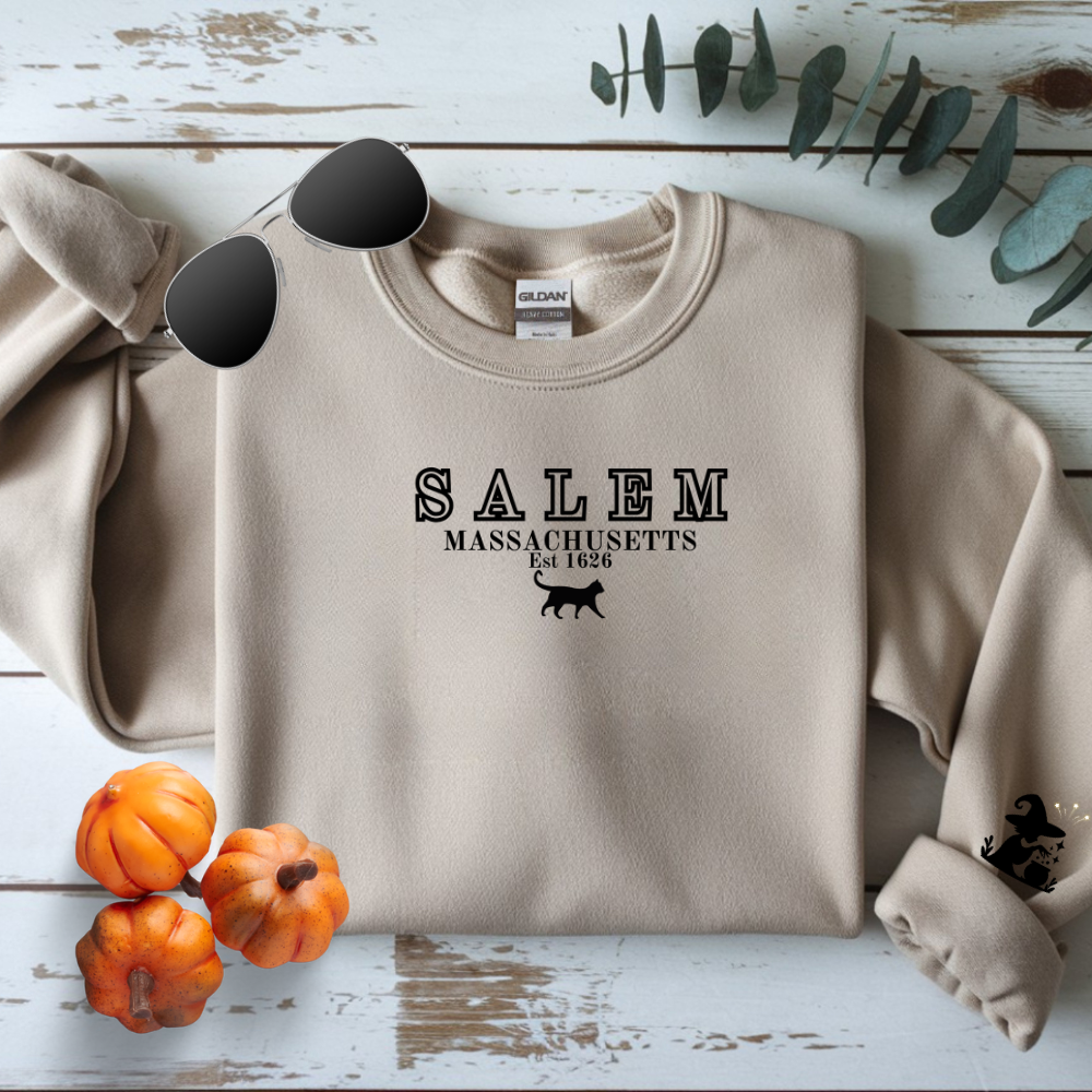 Beige sweatshirt with 'Salem, Massachusetts' text and graphics on a wooden surface with pumpkins and leaves.