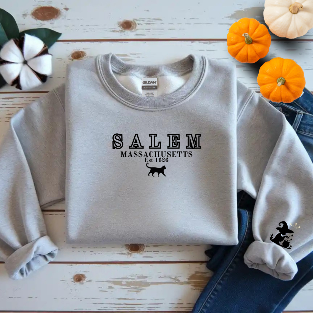 Gray sweatshirt with 'Salem Massachusetts Est. 1630' text and black cat graphic on a wooden surface with pumpkins.