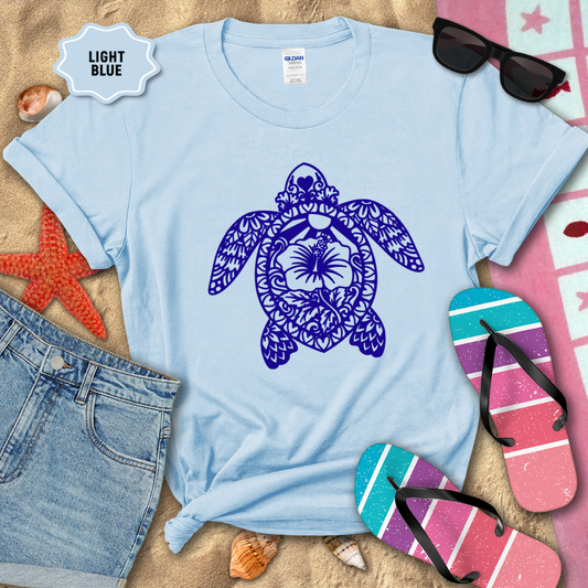 Light blue t-shirt with a purple turtle design on a sandy background with beach accessories.