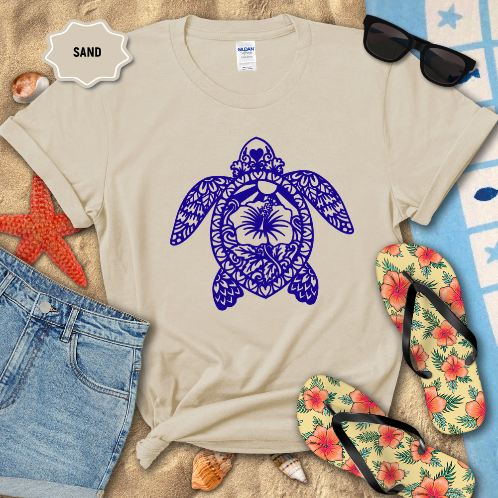 Beige t-shirt with blue turtle design on sand, surrounded by flip-flops and sunglasses.