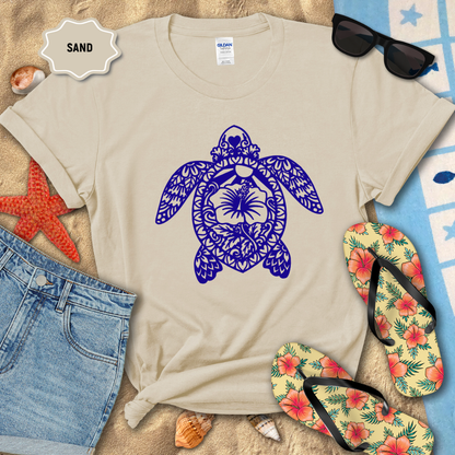 Beige t-shirt with blue turtle design on sand, surrounded by flip-flops and sunglasses.