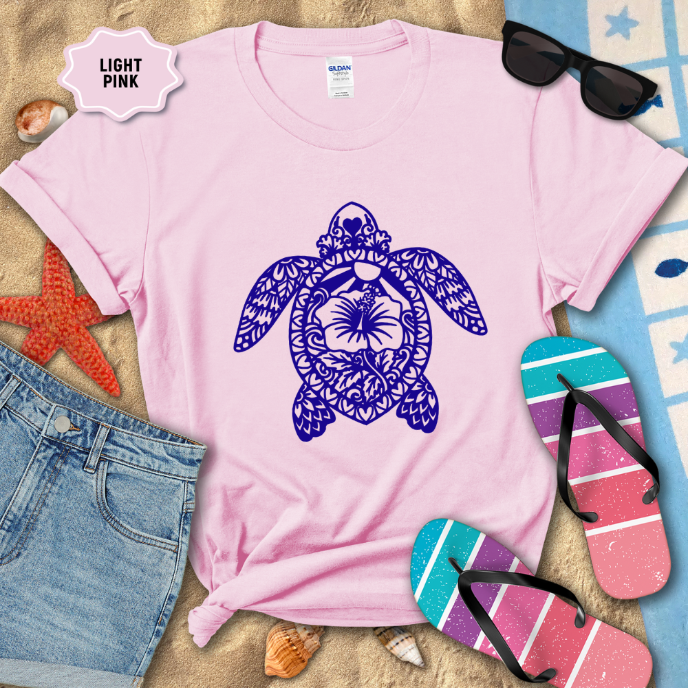 Light pink t-shirt with a blue turtle design on a sandy background with beach accessories.