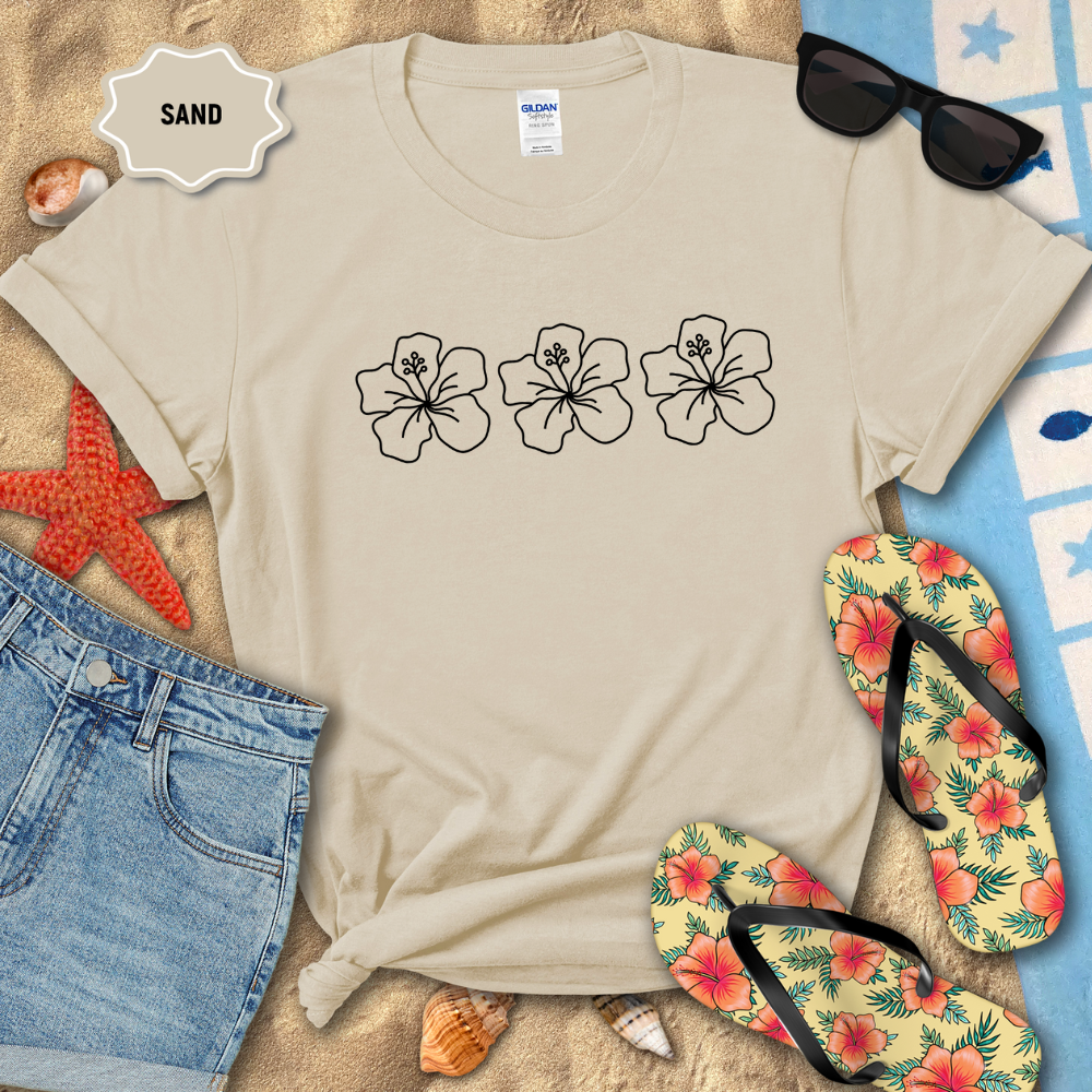 Beige t-shirt with floral designs on a sandy background with flip-flops and sunglasses.